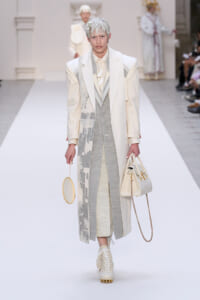 Runway model in a pale cream and gray layered suit with a long patterned coat, carrying a round handheld bag and a beige tote with a gold chain strap.
