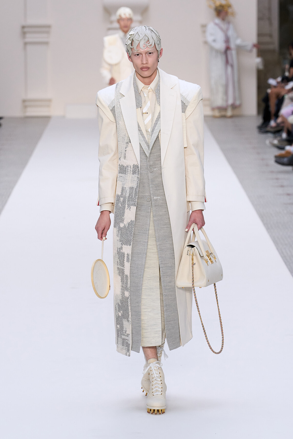 Runway model in a pale cream and gray layered suit with a long patterned coat, carrying a round handheld bag and a beige tote with a gold chain strap.