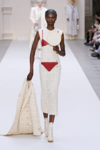 Model on a fashion runway wearing a white textured dress with red patches, holding a circular hoop with a delicate face sketch.