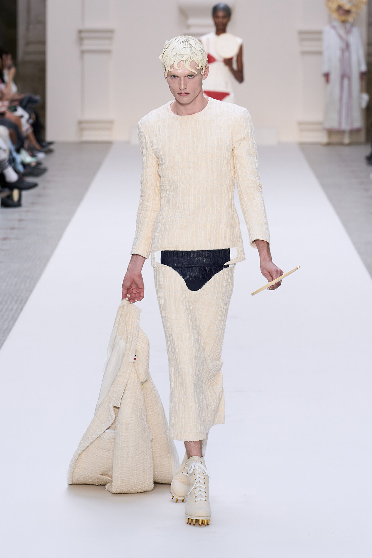 Male model walks a cream textured suit on a runway, holding a matching coat in one hand and a slim stick in the other, wearing a sculptural cream headpiece.