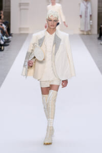 Model walks a white runway in an ivory suit with layered jacket, cream shirt, and shorts; she carries a small round handheld mirror and wears tall lace-up boots with gold studs.