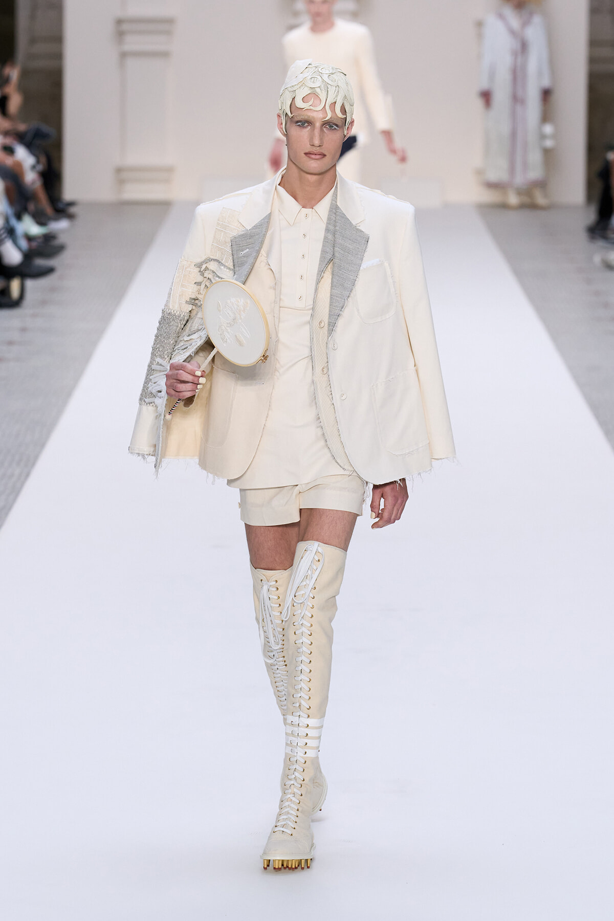 Model walks a white runway in an ivory suit with layered jacket, cream shirt, and shorts; she carries a small round handheld mirror and wears tall lace-up boots with gold studs.