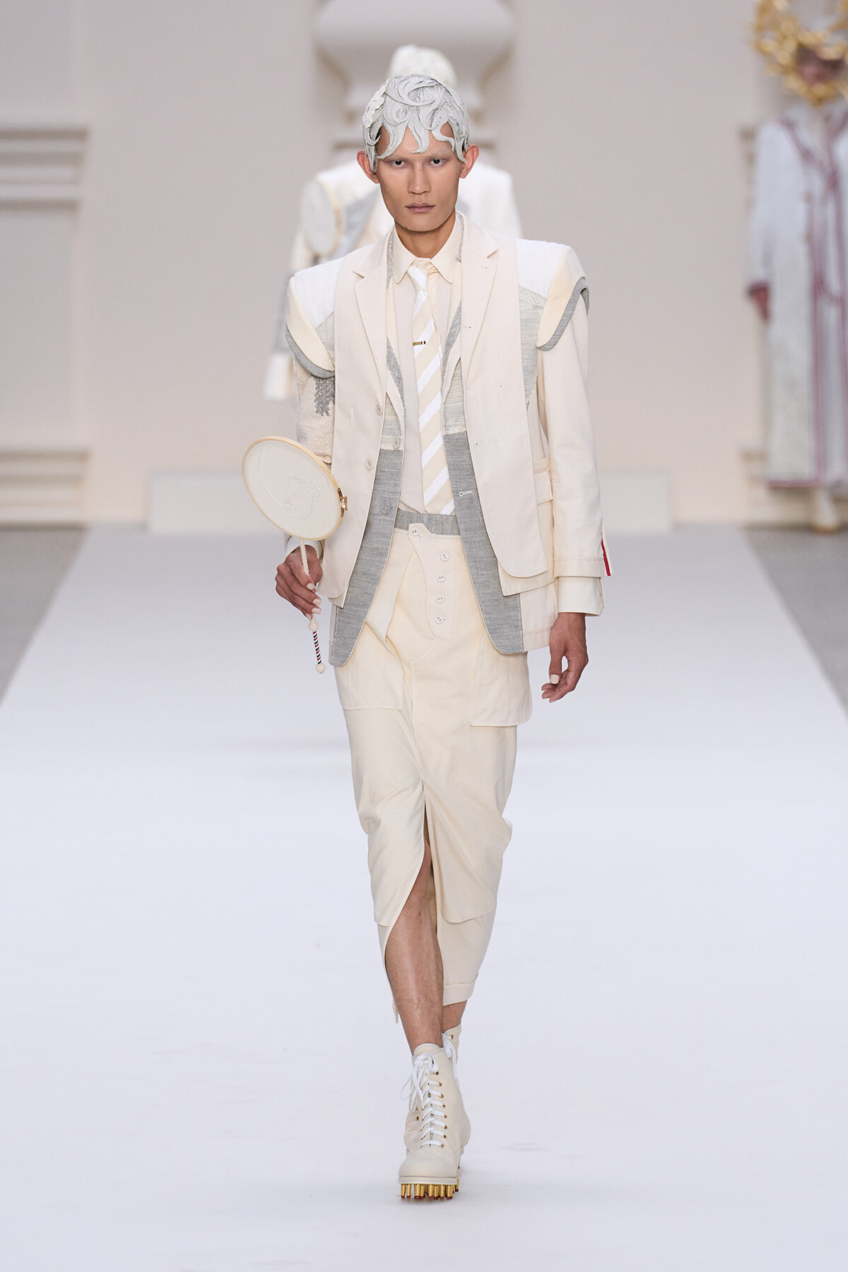 Male model in a cream suit with layered jacket and shorts walks a white runway, holding a round handheld mirror and wearing an intricate white headpiece.