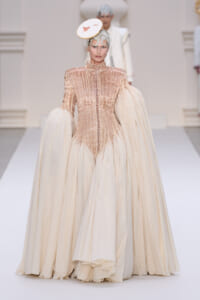 Model walks the runway in a champagne gown with a beaded bodice, high neck, and dramatic voluminous sleeves, topped with a small white circular hat.