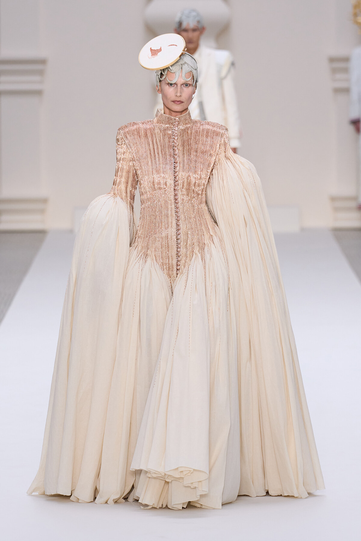 Model walks the runway in a champagne gown with a beaded bodice, high neck, and dramatic voluminous sleeves, topped with a small white circular hat.