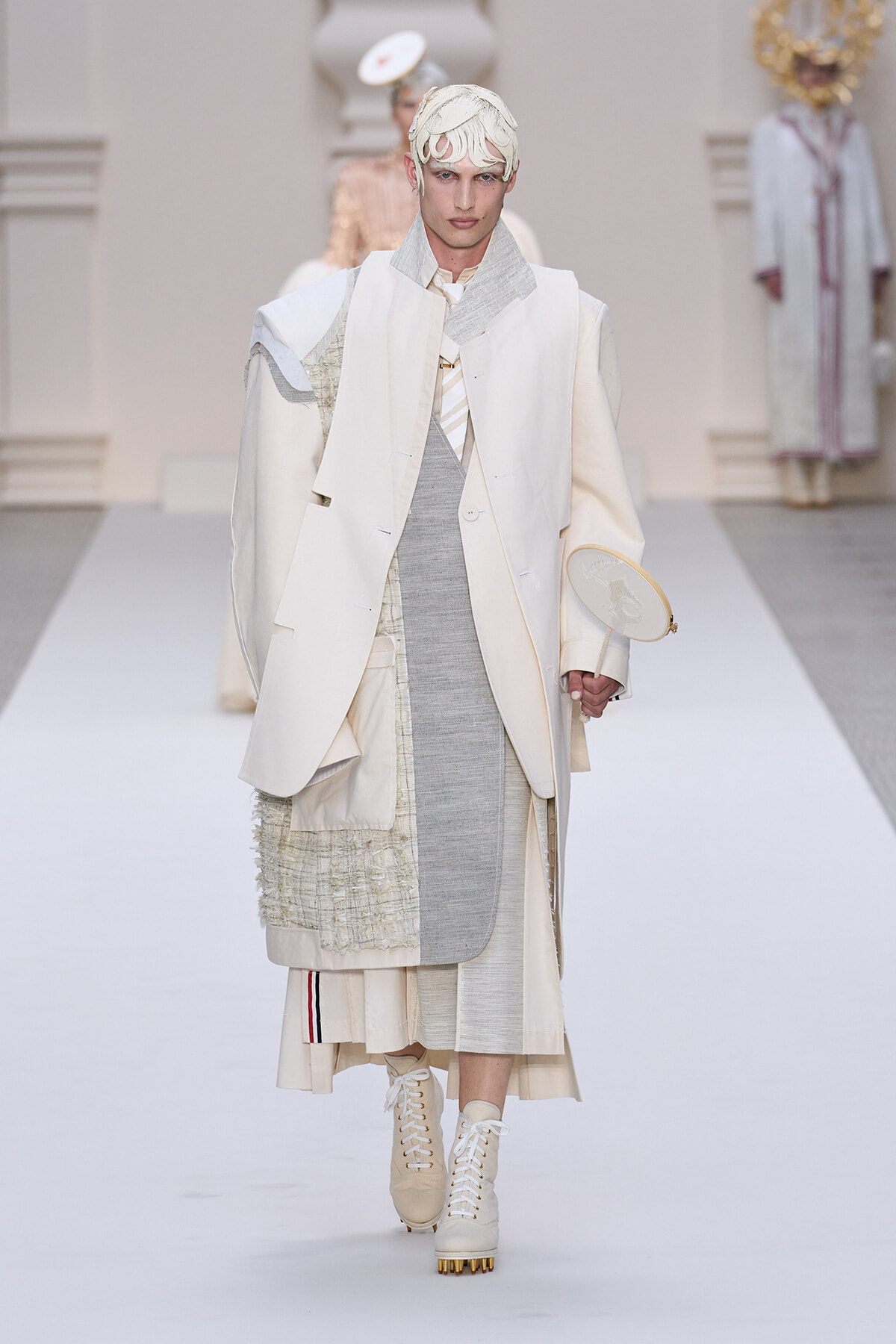 Male model walks a runway in an all-white layered outfit with a long cream coat, textured panels, and cream lace-up boots.