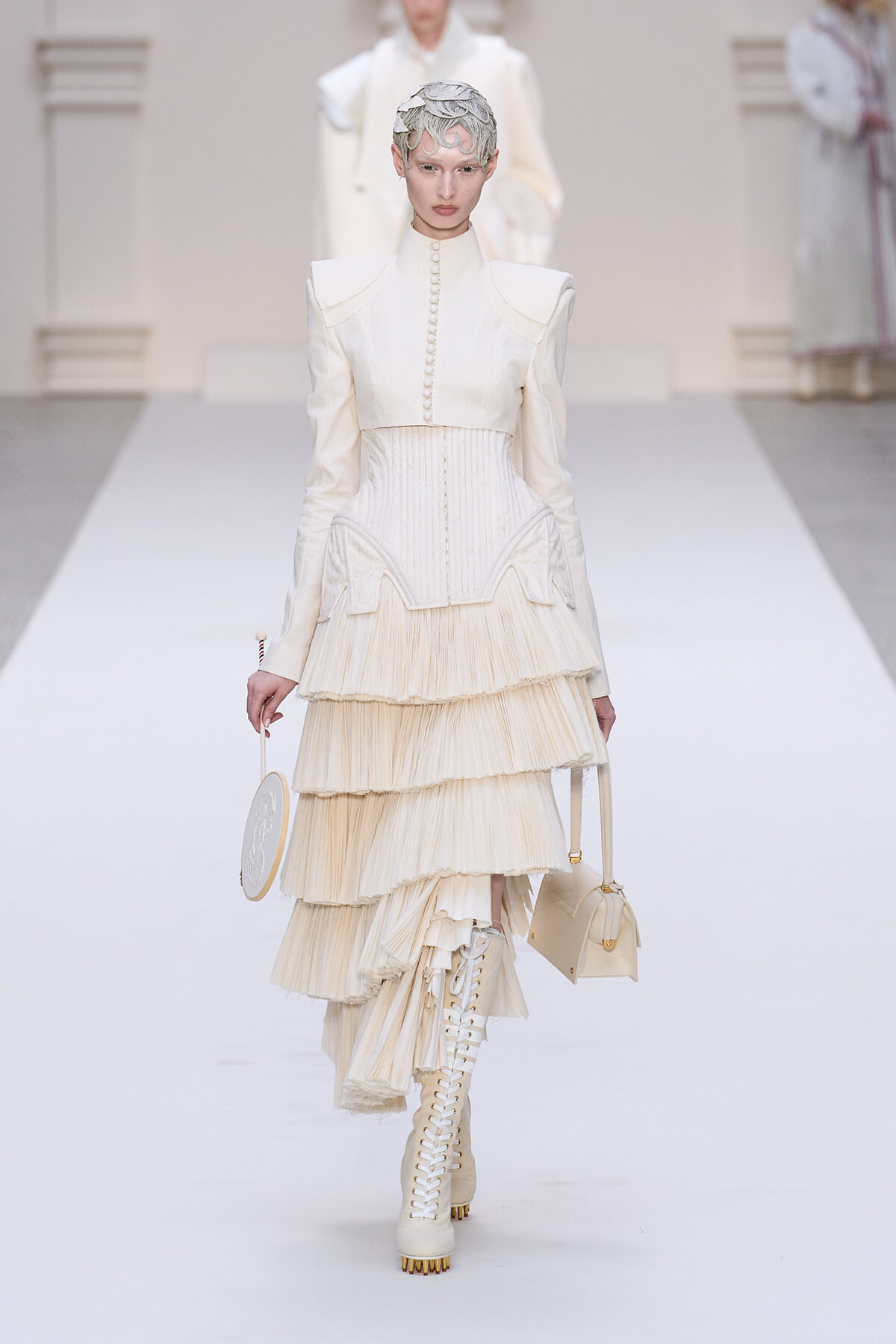 Model walking a cream ivory haute-couture gown with a structured bodice, pearl buttons, and a tiered pleated skirt, paired with lace-up boots and a round bag.