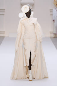 Model on a runway wearing an ivory sculptural gown with a flowing cape and a circular headpiece, white lace-up boots visible at the bottom.