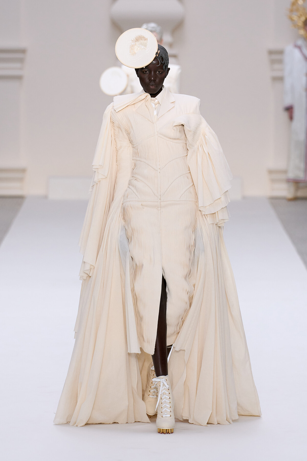 Model on a runway wearing an ivory sculptural gown with a flowing cape and a circular headpiece, white lace-up boots visible at the bottom.