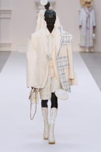 Model walks the runway in a cream-toned layered suit with a textured patchwork jacket and knee-high lace-up boots, carrying a small beige handbag with a chain strap.