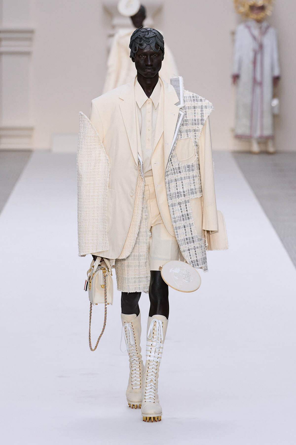 Model walks the runway in a cream-toned layered suit with a textured patchwork jacket and knee-high lace-up boots, carrying a small beige handbag with a chain strap.
