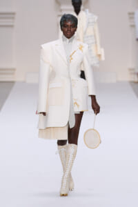Model on a white runway wearing an oversized cream blazer with gold embroidery, a light gray top, a pleated hem, knee-high lace-up boots, and a circular embroidery bag.