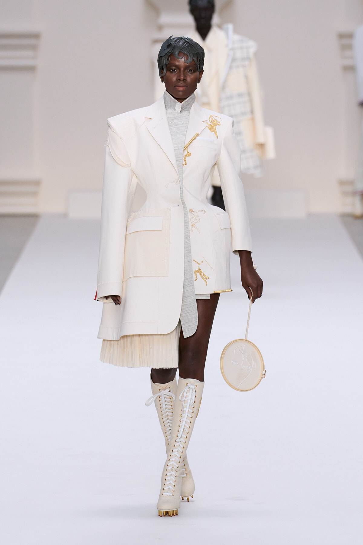 Model on a white runway wearing an oversized cream blazer with gold embroidery, a light gray top, a pleated hem, knee-high lace-up boots, and a circular embroidery bag.