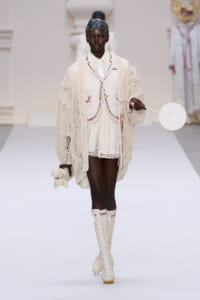 Black model walks a runway in a cream knit coat over matching shorts with red/black piping, tall lace-up boots, a headscarf, and holds a round white handbag with a small plush toy.