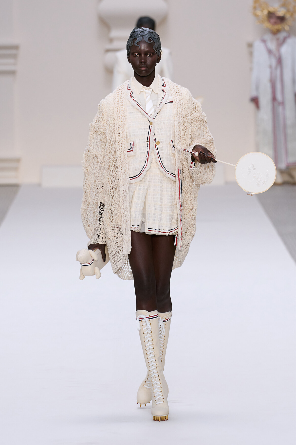 Black model walks a runway in a cream knit coat over matching shorts with red/black piping, tall lace-up boots, a headscarf, and holds a round white handbag with a small plush toy.