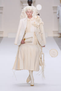 Runway model in a cream ensemble with a tailored blazer over a pleated beige dress, floral appliqués, clutch in hand and a circular embroidery hoop prop.