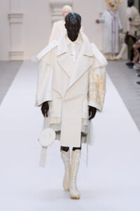 Model on a white runway wearing an oversized cream coat, matching shirt, knee-high lace-up boots, and a circular white handbag.