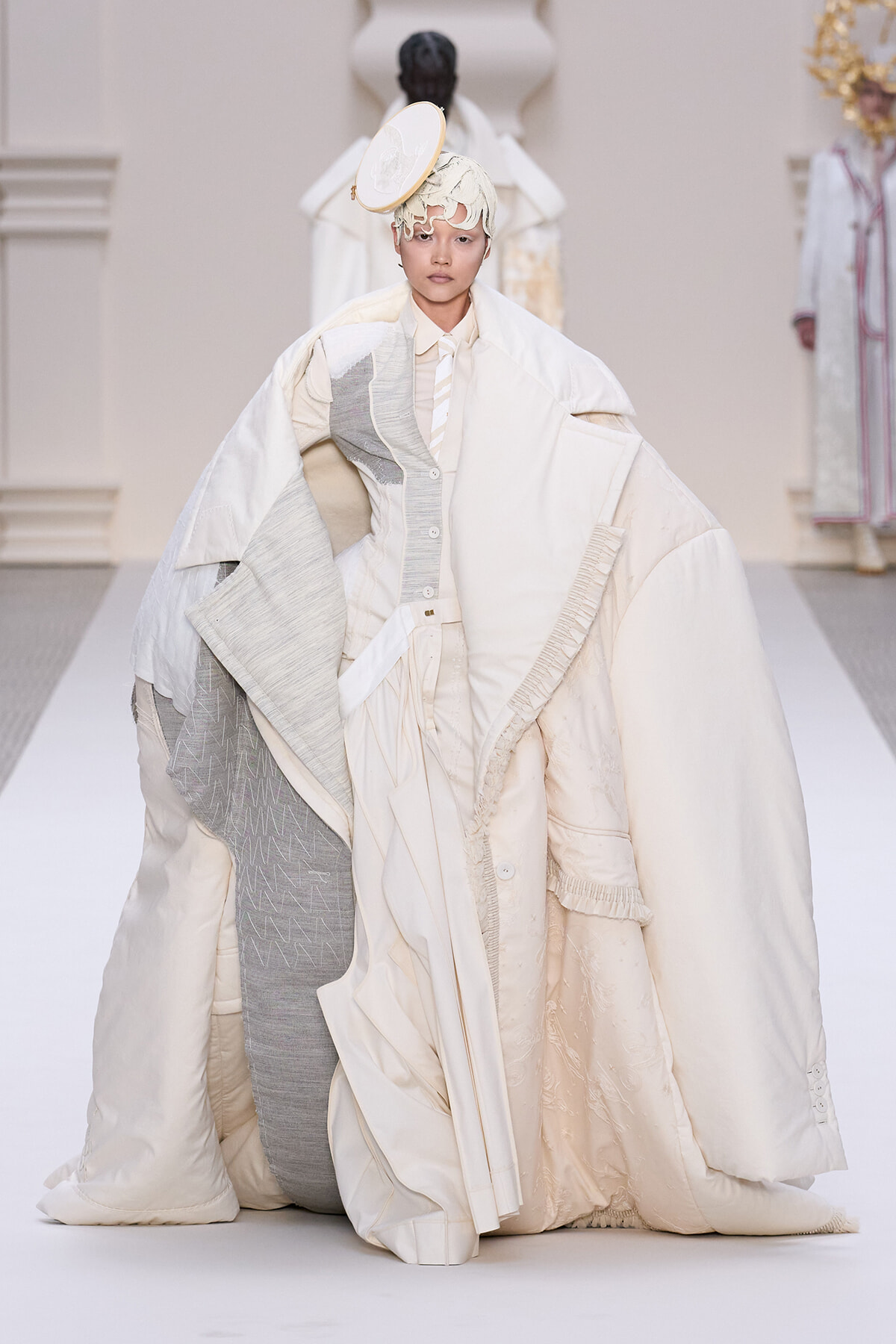 Model on a runway wearing an oversized cream quilted coat with layered panels and neutral-toned tailoring, creating a sculptural silhouette.
