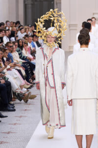 Model on a fashion show runway wearing a white sequined suit with red and blue piping and a large gold leaf headdress.