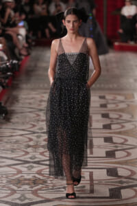 Model walks the runway in a black sheer midi dress with sparkling beads and a beaded bodice, wearing a large black bow in her hair.