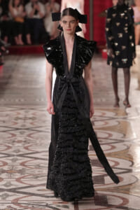 Model in a black avant-garde gown with oversized glossy ruffle shoulders, a waist tie, and a tiered, ruffled skirt on a fashion-show runway.