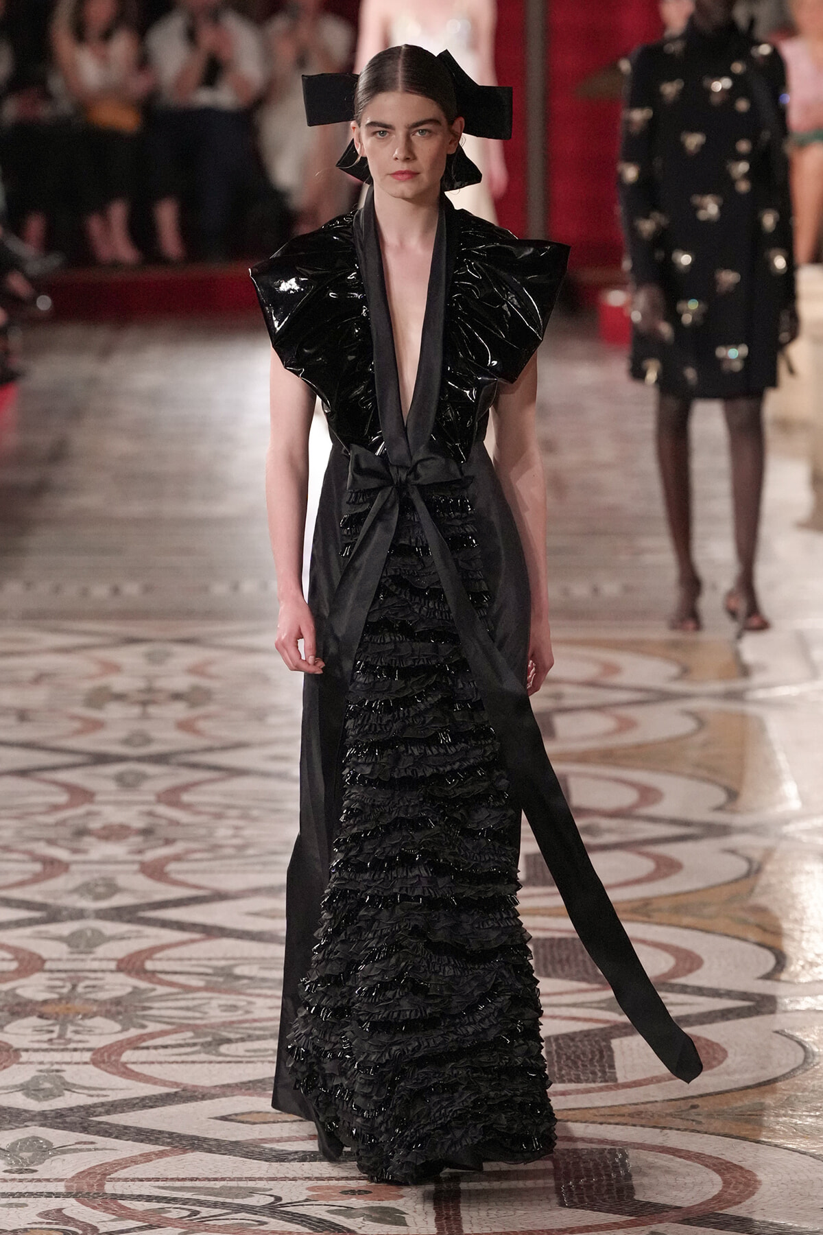 Model in a black avant-garde gown with oversized glossy ruffle shoulders, a waist tie, and a tiered, ruffled skirt on a fashion-show runway.