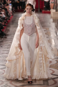 Model on a runway wearing a cream satin gown with a voluminous ruffled cape and a beaded pale pink corset top.