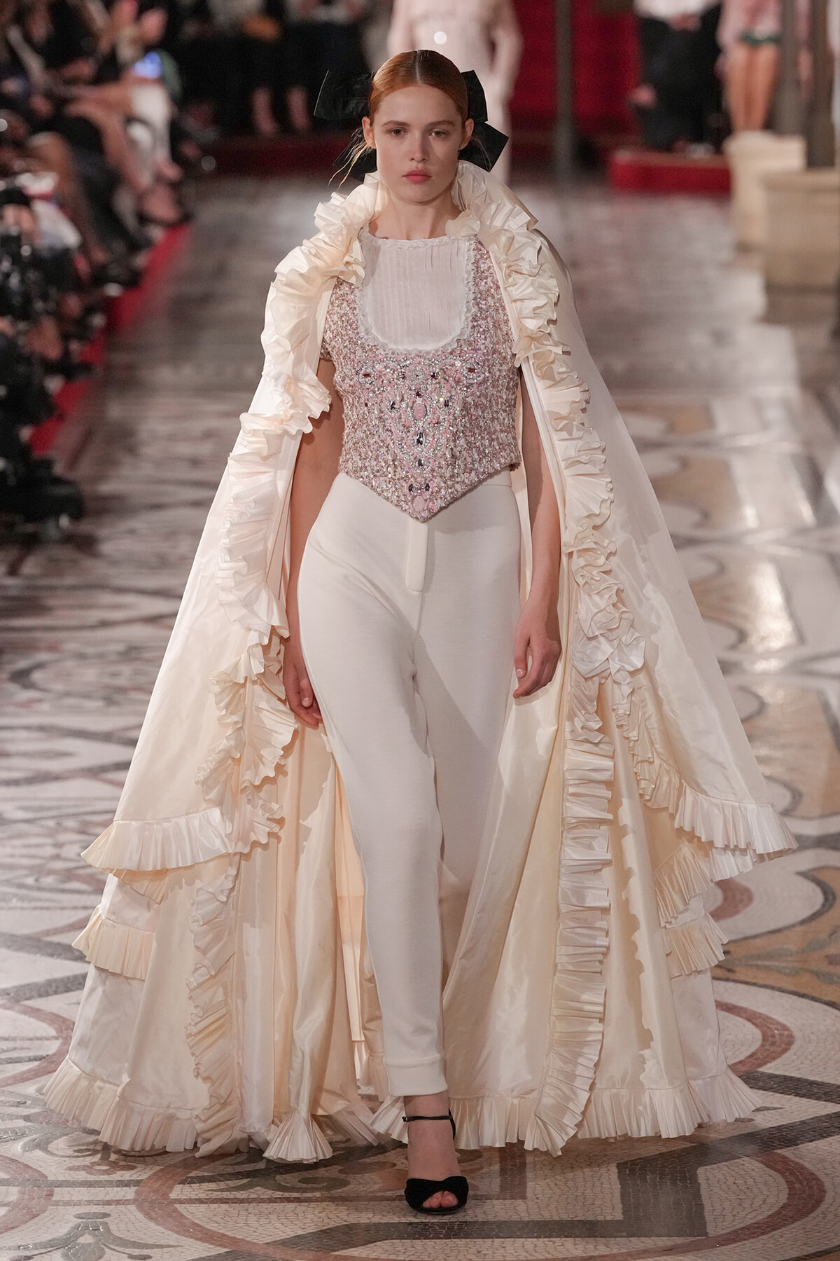 Model on a runway wearing a cream satin gown with a voluminous ruffled cape and a beaded pale pink corset top.
