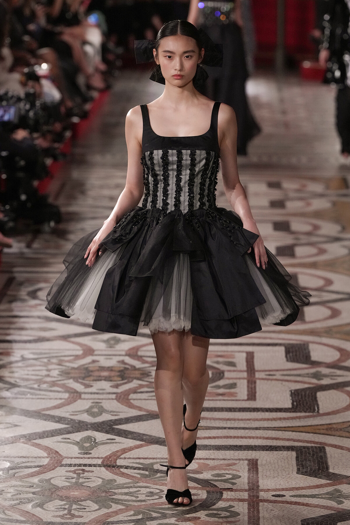 Model walking a runway in a black and gray corseted mini dress with a layered tulle skirt.