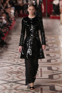 Model walks a runway in a glossy black patent coat with metal buttons, paired with sheer studded pants and black sandals.