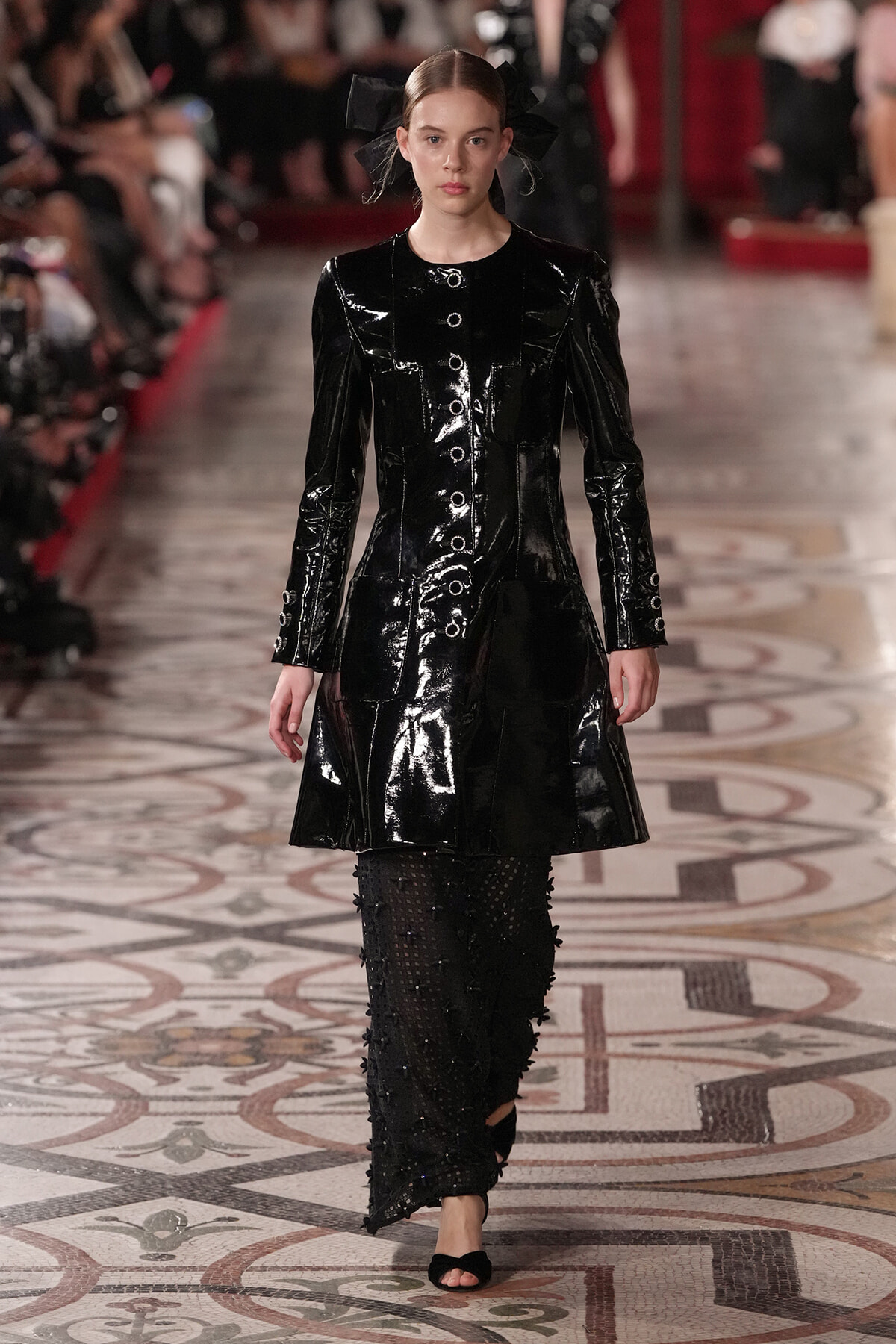 Model walks a runway in a glossy black patent coat with metal buttons, paired with sheer studded pants and black sandals.