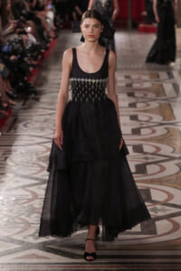 Runway model in a black gown with a beaded, embellished bodice and layered sheer skirt, walking on a patterned marble runway with a large black bow in her hair.