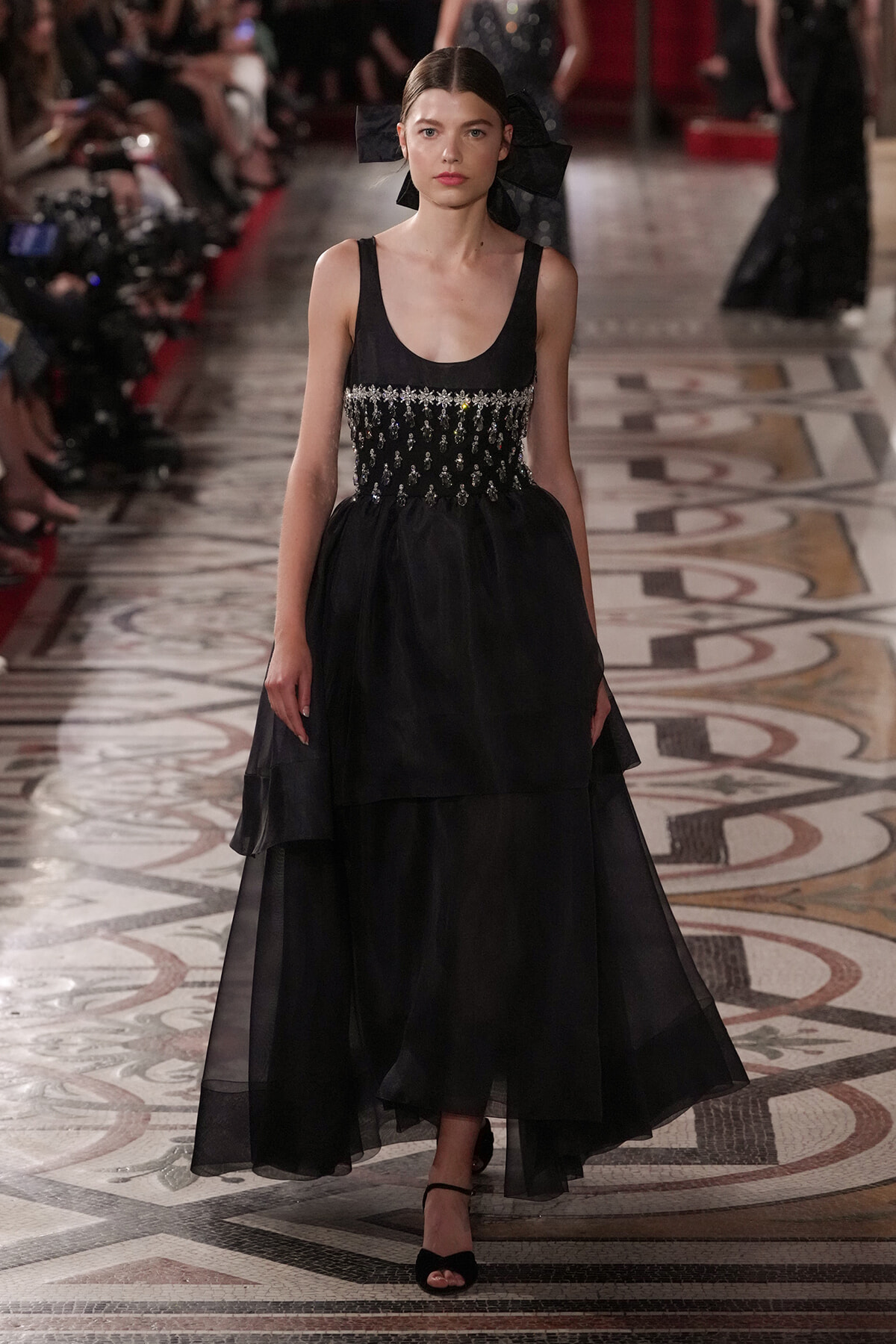 Runway model in a black gown with a beaded, embellished bodice and layered sheer skirt, walking on a patterned marble runway with a large black bow in her hair.