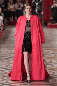 Model on a runway wearing a bright pink full-length cape over a black lace top and shiny black mini skirt, with black sandals and a large black bow in her hair.