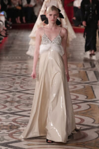 Runway model in a pale cream satin gown with a jeweled bodice and feathered waist, her hair tied with a large black bow.
