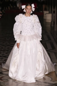 Model on a runway wearing a white ballgown with voluminous puff sleeves, a floral-embellished bodice, and a full, structured skirt.