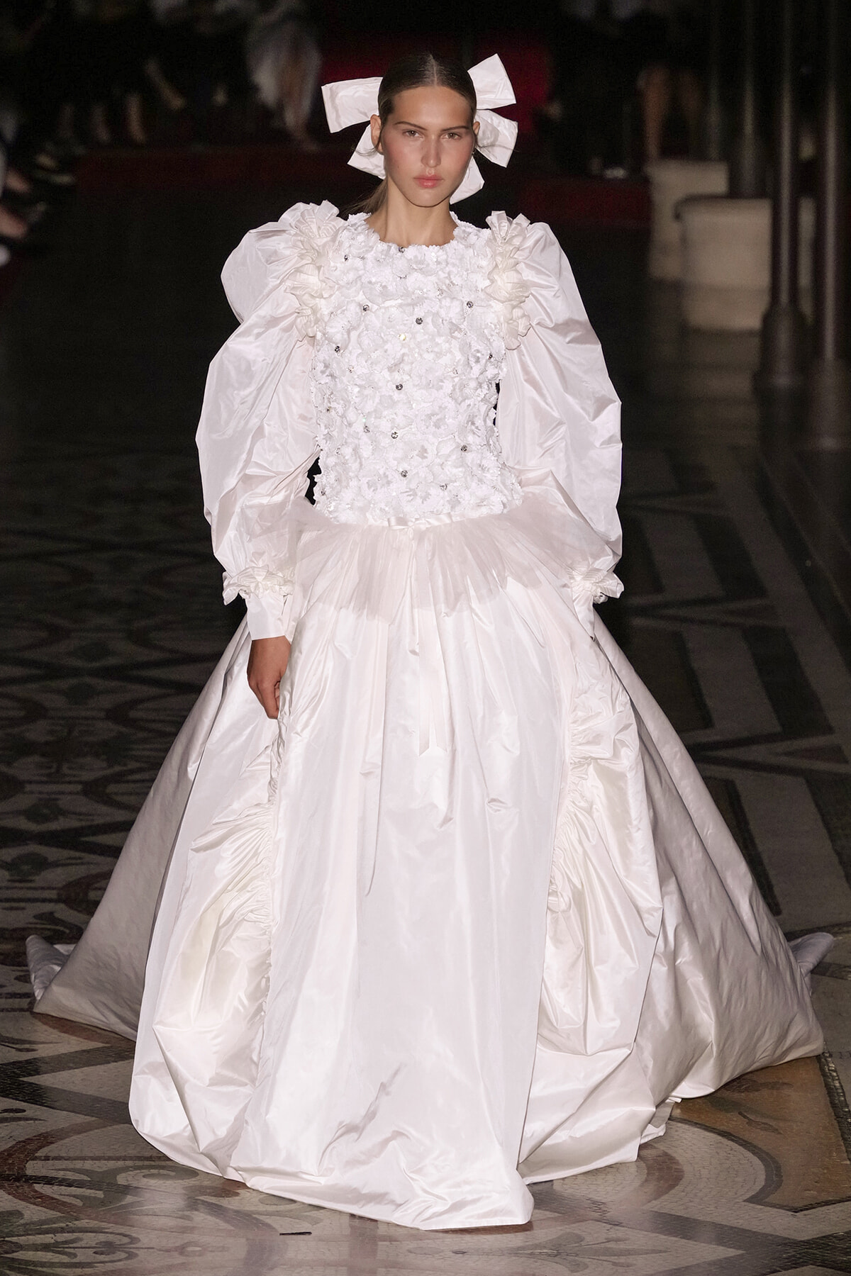 Model on a runway wearing a white ballgown with voluminous puff sleeves, a floral-embellished bodice, and a full, structured skirt.