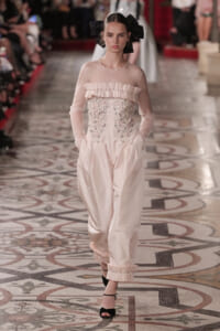 Model walks the runway in a pale pink, sheer-sleeve gown with a jeweled, ruffled bodice and pockets, topped by a large black bow in hair.