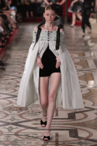 Model walks the runway in a pale blue off‑the‑shoulder gown with an embroidered bodice and long cape over a black mini dress, paired with black high heels and ribbon accents in the hair.