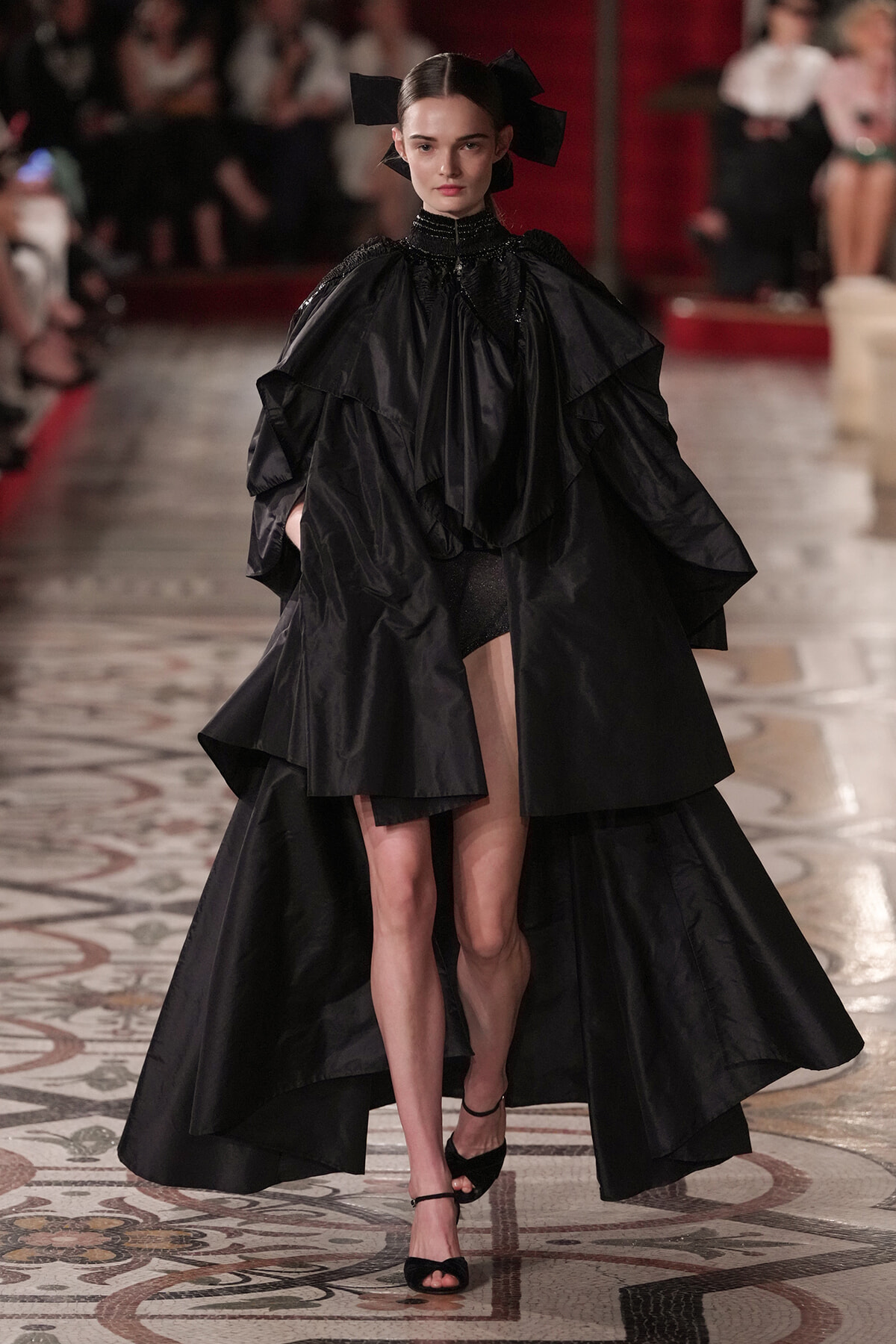Model walks the runway in a dramatic black cape gown with layered ruffles and a high collar, wearing black heels and a bow headpiece.