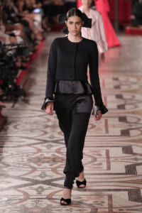 Model walks the runway in a black tailored jacket with peplum and matching pants, accessorized with a large black bow headpiece and black open-toe heels.