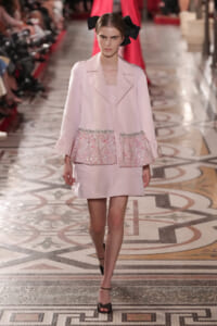 Model walking a runway in a pale pink coat with sparkly sequin waist panels and a large black bow in her hair.