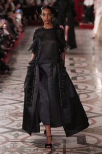 Runway model in a black satin gown with cascading panels, beaded trims along the seams, and sheer puff sleeves, walking on a patterned catwalk.