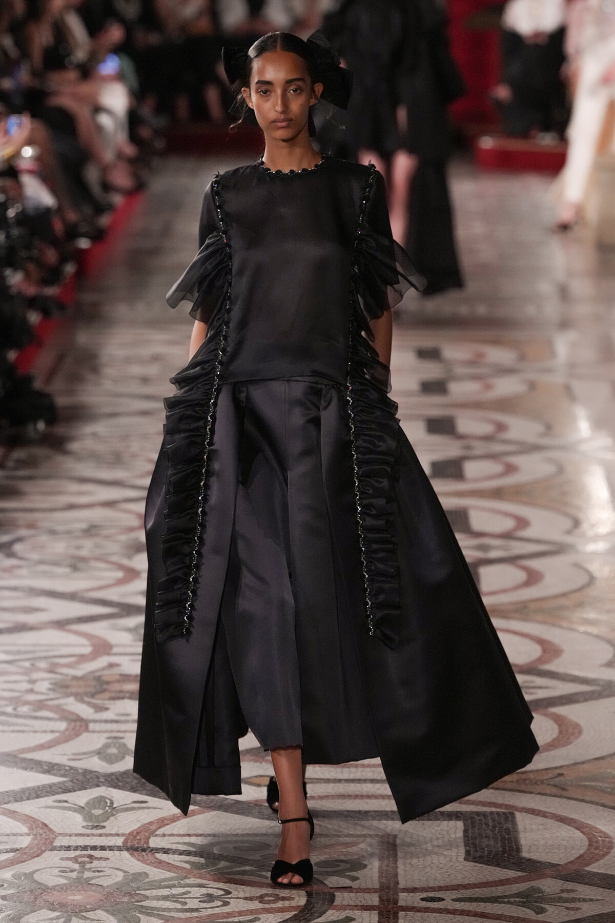 Runway model in a black satin gown with cascading panels, beaded trims along the seams, and sheer puff sleeves, walking on a patterned catwalk.