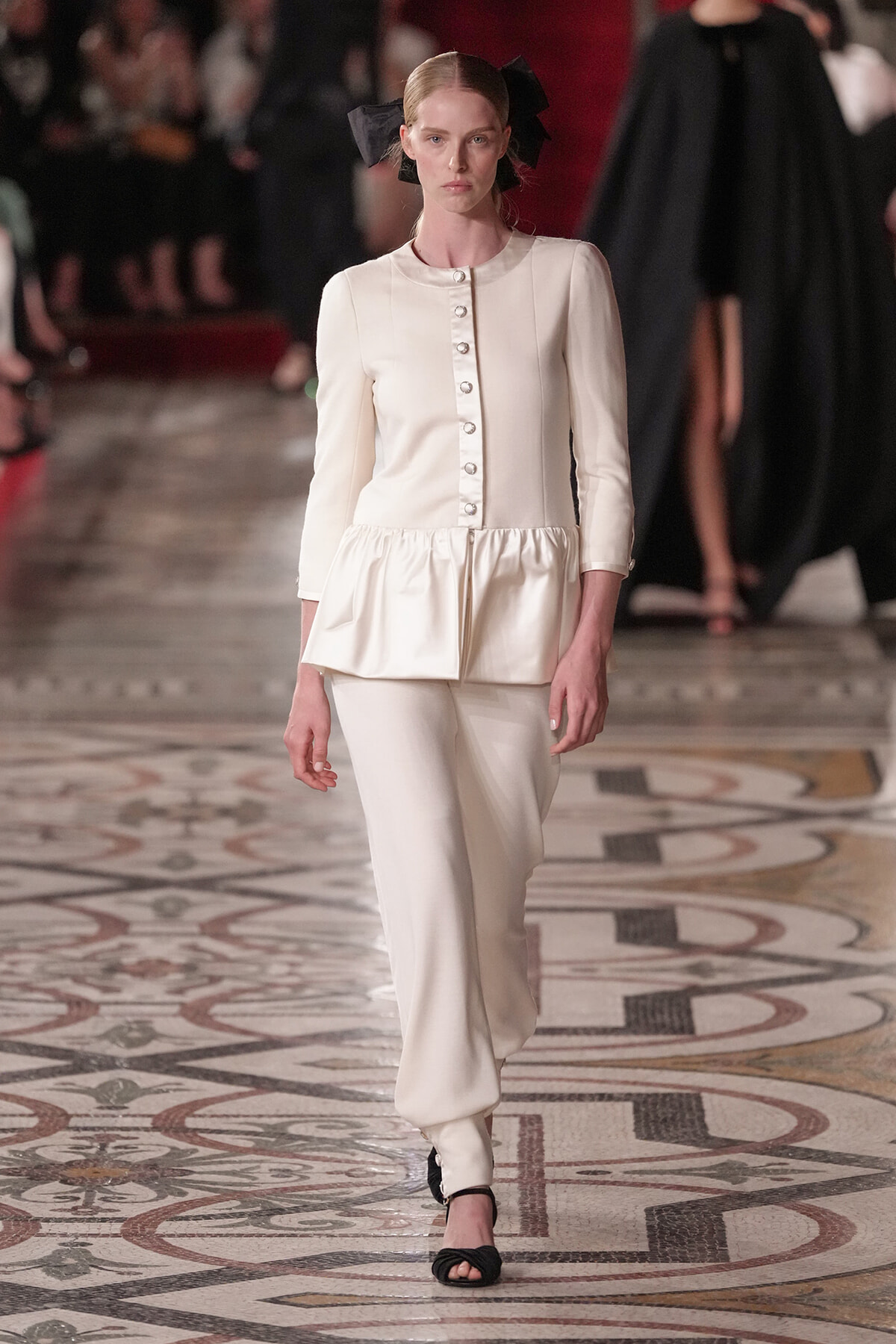 Model walks the runway in an ivory peplum-buttoned top with matching wide-leg pants, black heeled sandals, and a black bow in hair.