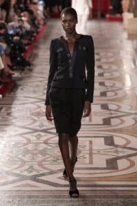 Model walking a fashion runway in a black structured blazer and knee-length skirt set.