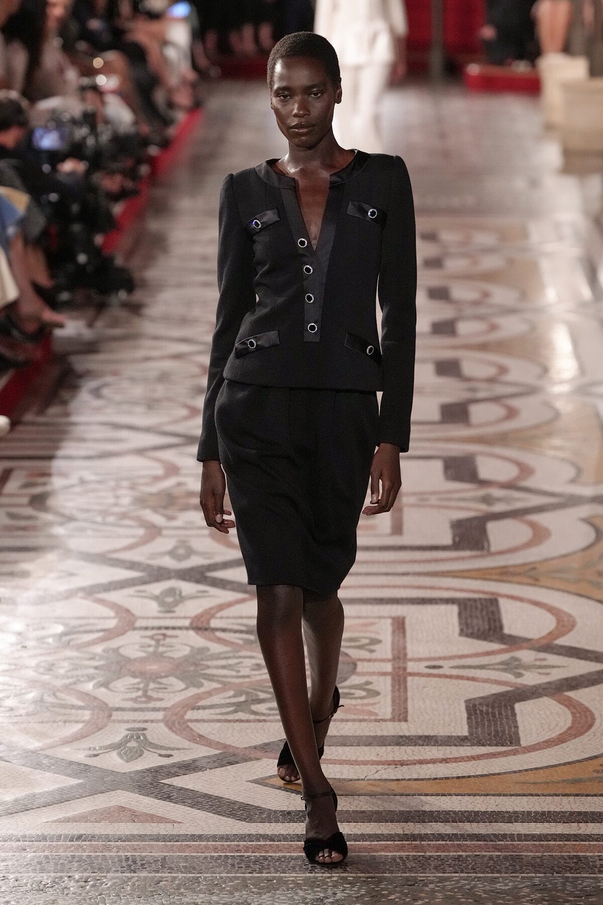 Model walking a fashion runway in a black structured blazer and knee-length skirt set.