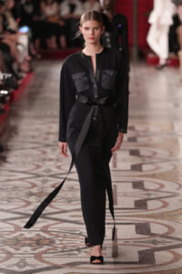 Model walks a fashion runway in a black belted jumpsuit with front pockets and a deep neckline, wearing black heeled sandals.