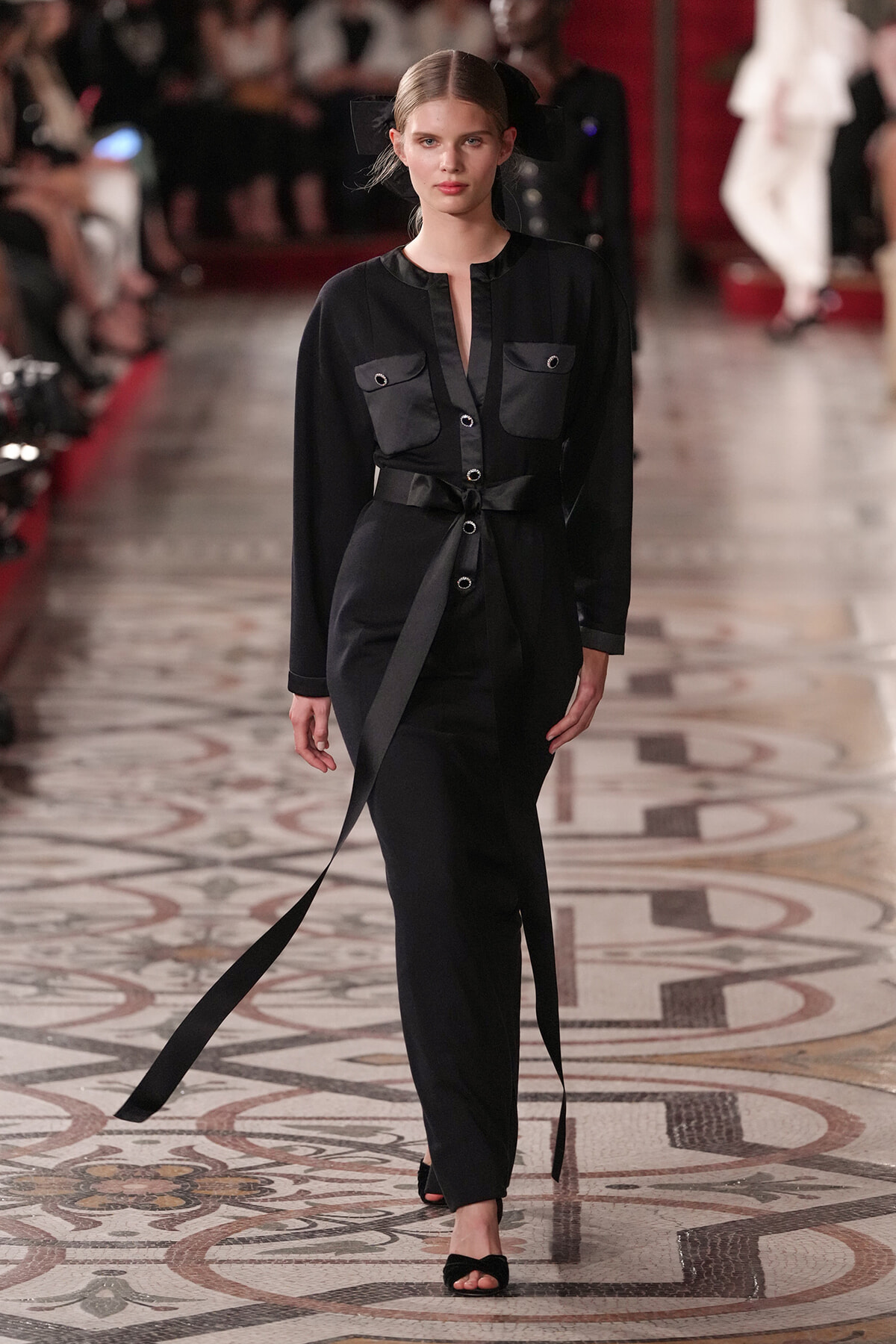 Model walks a fashion runway in a black belted jumpsuit with front pockets and a deep neckline, wearing black heeled sandals.