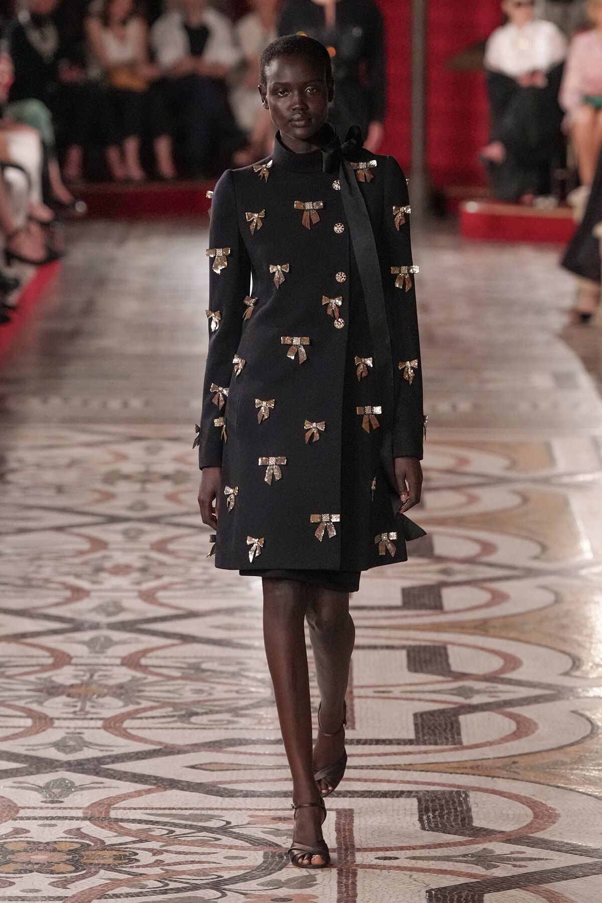 Black knee-length coat with gold bow appliques on a runway, model walking in heels. Context: fashion show.